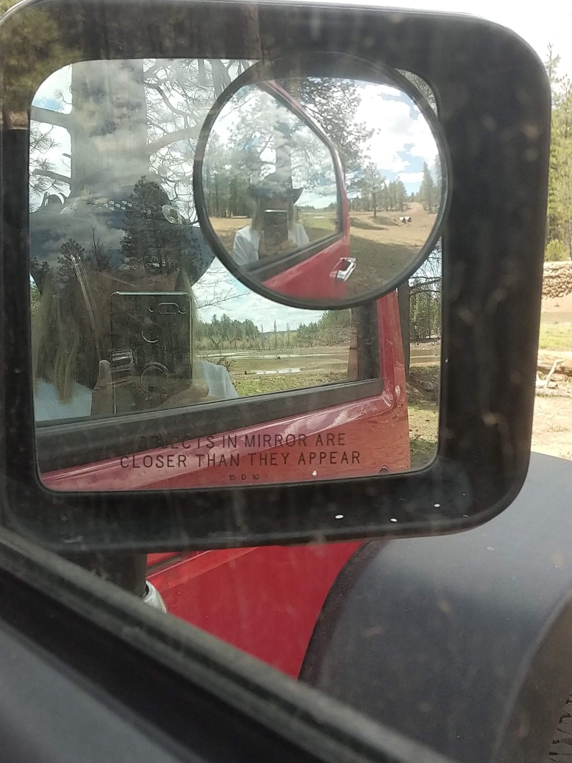 Jo photographing a wooded travel scene, reflected in a friend’s Jeep mirror beside the words Objects in mirror are closer than they appear